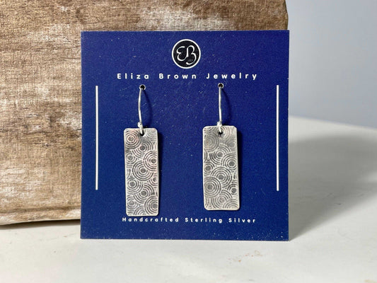 Elegant circle patterned silver earrings on blue card, handcrafted in sterling silver with a textured patina finish, ideal for casual or formal wear.